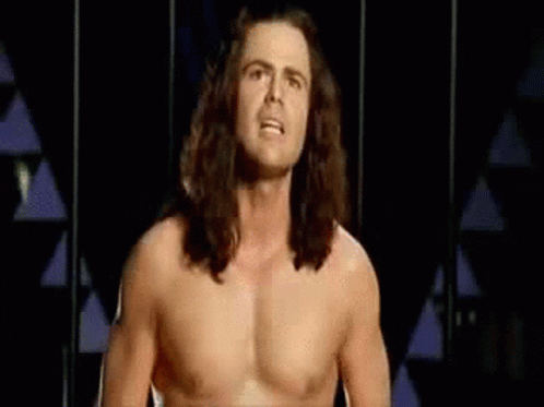 Shirtless Man With Long Hair Standing in Front of Triangle Wall