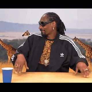 Snoop Dogg Is Sitting At A Table With A Blue Cup And Giraffes In The Background . GIF