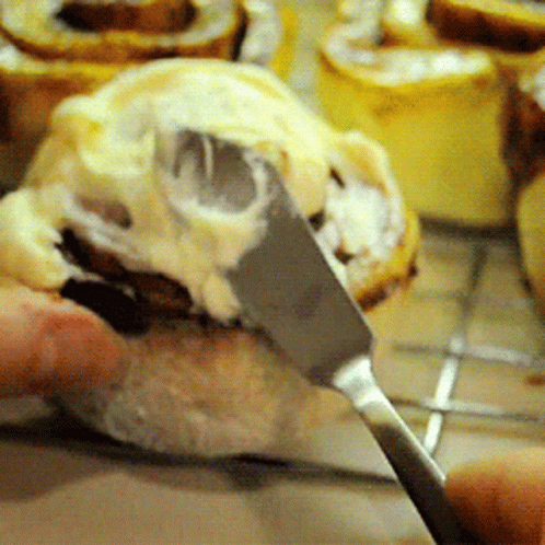 Someone Is Spreading Frosting On A Cinnamon Roll With A Knife GIF
