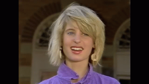 The Woman Is Wearing A Purple Shirt And Earrings And Is Smiling . GIF