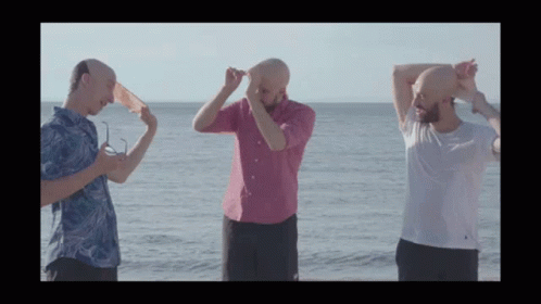 Three Bald Men Are Standing On The Beach With Their Heads In The Air GIF