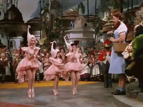 Three Ballerinas In Pink Tutus Are Dancing In Front Of A Crowd While A Woman In A Blue Dress Stands Behind Them . GIF