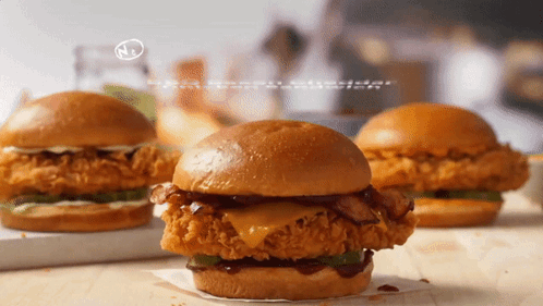 Three Fried Chicken Sandwiches On A Wooden Table With A N On The Bottom Right GIF