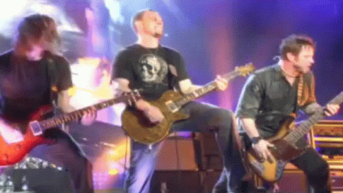Three Men Are Playing Guitars On A Stage And One Of Them Is Wearing A Black Shirt With A Skull On It GIF