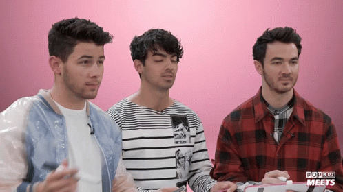 Three Men Are Sitting In Front Of A Pink Background That Says Popbuzz Meets On It GIF