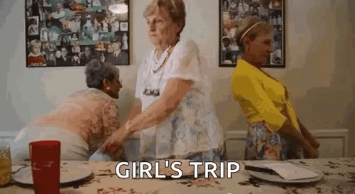 Three Older Women Are Standing Around A Table With The Words `` Girl 'S Trip '' Above Them . GIF