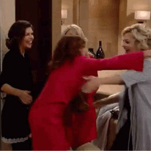 Three Women Are Hugging Each Other In A Room . One Of The Women Is Wearing A Red Dress . GIF