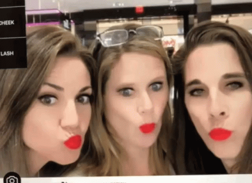 Three Women Are Making Funny Faces In Front Of A Sign That Says Cheek Lash And Lipstick GIF