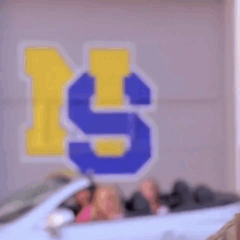 Three Women Are Sitting In A White Convertible Car In Front Of A Wall With The Letter S Painted On It . GIF