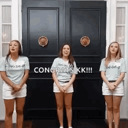 Three Women Are Standing In Front Of A Door With The Words `` Congrats Kk '' Written On It . GIF