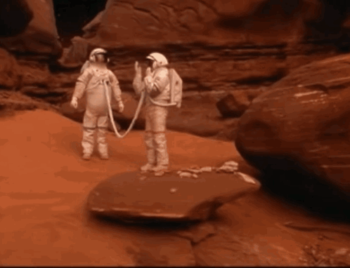 Two Astronauts Standing Next To Each Other On A Rock In The Desert GIF