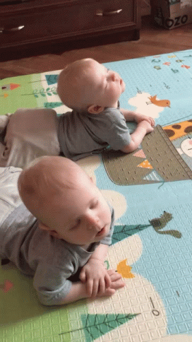 Two Babies Laying On A Mat With A Giraffe On It And A Box That Says Ozo On It GIF