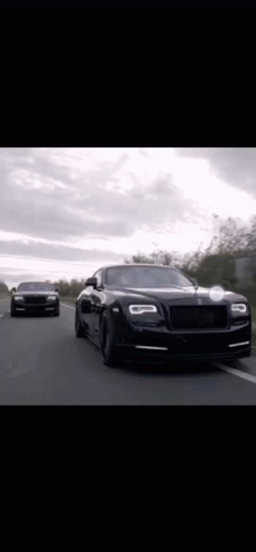 Two Black Rolls Royce Cars Are Driving Down A Road . GIF