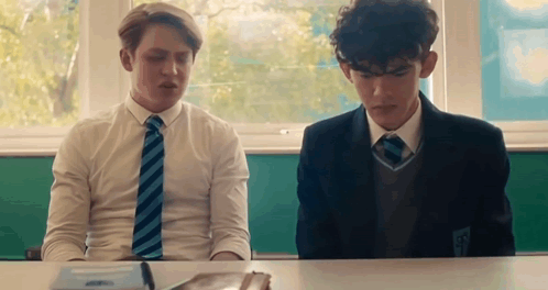 Two Boys Are Sitting At A Desk In A Classroom . GIF