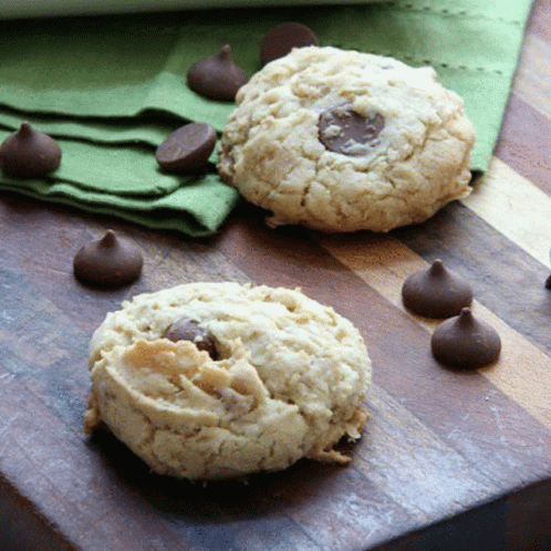 Two Cookies On A Wooden Cutting Board With Chocolate Chips Around Them GIF