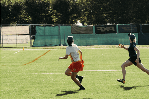Two Female Athletes Running on Field with BKF Banner
