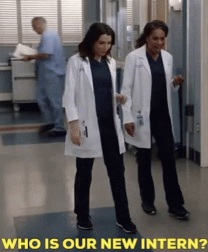 Two Female Doctors Are Walking Down A Hallway With The Words " Who Is Our New Intern " Behind Them GIF