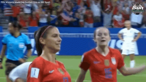 Two Female Soccer Players On A Field With A Referee In The Background GIF