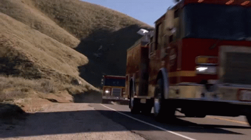 Two Fire Trucks Are Driving Down A Road With A Mountain In The Background GIF