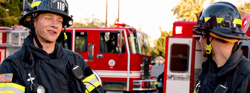 Two Firefighters Are Talking In Front Of A Fire Truck With The Number 118 On The Helmet GIF