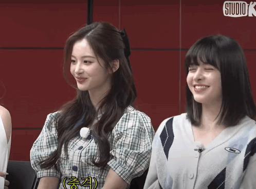 Two Girls Are Sitting Next To Each Other And Smiling In Front Of A Red Wall That Says Studio K GIF