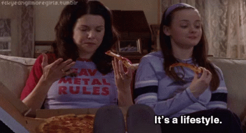 Two Girls Are Sitting On A Couch Eating Pizza And One Has A Shirt That Says " Raw Metal Rules " GIF