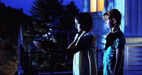 two girls are standing in front of a house at night and one is holding a bowl