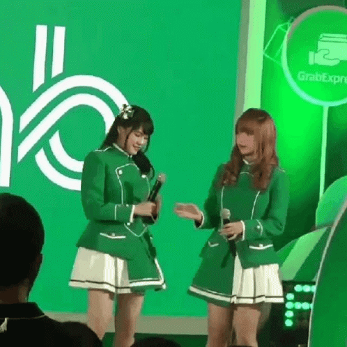 Two Girls In Green Uniforms Are Standing Next To Each Other With Microphones In Front Of A Green Background . GIF