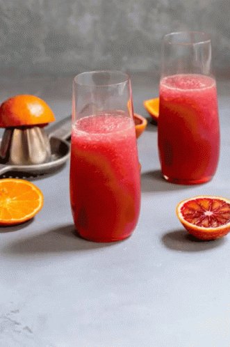 Two Glasses Of Red Juice Next To Oranges And A Juice Press GIF