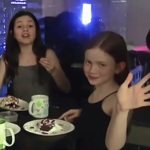 Two Little Girls Are Sitting At A Table With Plates Of Food GIF