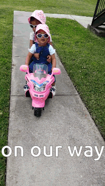 Two Little Girls Riding A Pink Toy Motorcycle On A Sidewalk With The Words On Our Way Below Them GIF