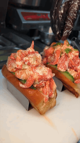 Two Lobster Rolls Are Being Prepared On A Cutting Board By A Person Wearing Black Gloves GIF
