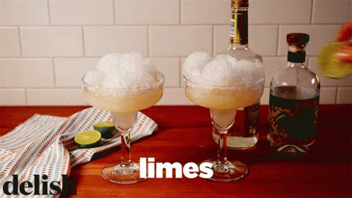 Two Margarita Glasses Filled With Ice And Limes Next To A Bottle Of Liquor GIF