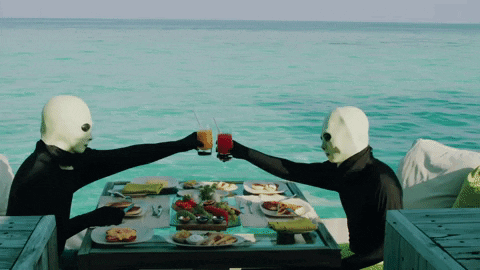 Two Masked People Toasting At A Table With Food GIF