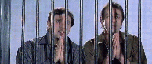 Two Men Are Behind Bars With Their Hands Folded In Front Of Them GIF