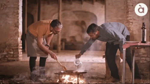 Two Men Are Cooking Over A Fire With The Letter A On The Bottom Right GIF