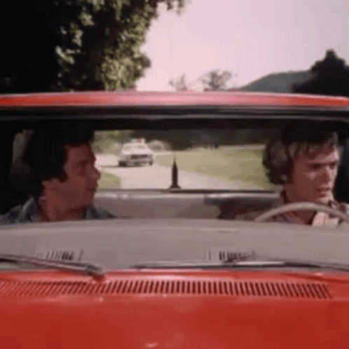 Two Men Are Driving A Red Car Down A Road . GIF