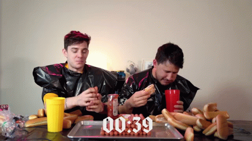 Two Men Are Eating Hot Dogs And Drinking Soda In Front Of A Tray Of Hot Dogs That Says 00:39 On It GIF