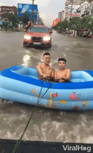 Two Men Are In An Inflatable Pool In A Flooded Street With A Car Behind Them GIF