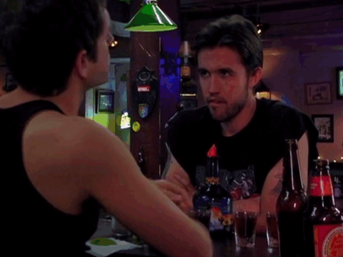 Two Men Are Sitting At A Bar With Bottles Of Beer And One Has A Sticker That Says ' Coca Cola ' On It GIF