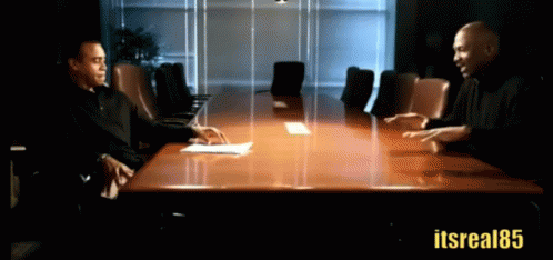 Two Men Are Sitting At A Conference Table With The Hashtag Itsreal85 GIF