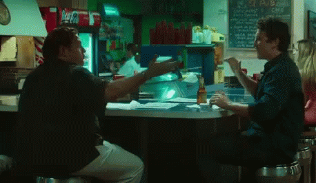 two men are sitting at a counter in a restaurant with a coca cola fridge in the background
