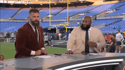 Two Men Are Sitting At A Desk In Front Of A Stadium That Says M & T Bank Stadium GIF