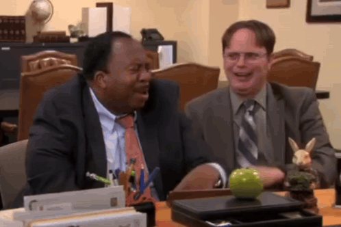 Two Men Are Sitting At A Desk With A Green Apple On It GIF