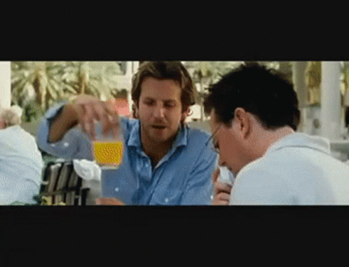 Two Men Are Sitting At A Table And One Is Pouring Orange Juice Into Another Man 'S Glass GIF
