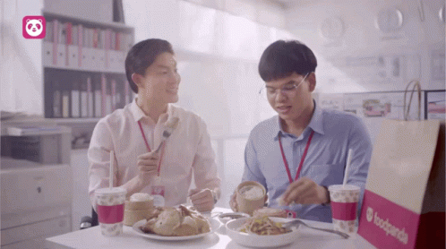 Two Men Are Sitting At A Table Eating Food And Drinking From A Bag That Says Foodpanda GIF