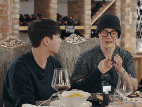 Two Men Are Sitting At A Table Eating Food And Drinking Wine With Bottles Of Wine In The Background GIF