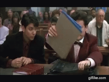 Two Men Are Sitting At A Table In A Courtroom And One Of Them Is Holding A Folder GIF