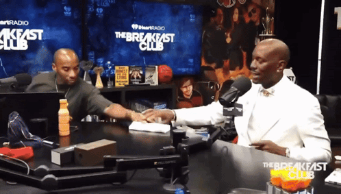 Two Men Are Sitting At A Table In Front Of A Sign That Says The Breakfast Club GIF