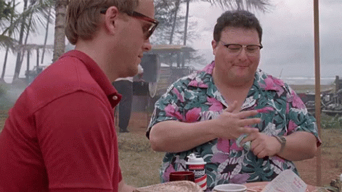two men are sitting at a table talking to each other . one of the men is wearing sunglasses .